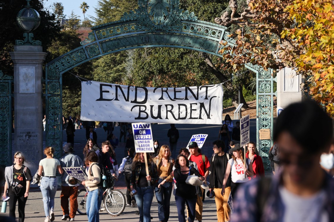 People gather with UAW on Strike/Unfair Labor Practice signs at a university gate, over which is hung a large banner that reads END RENT BURDEN